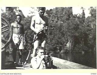 An unidentified native man looks on while NX170988 Spr R Skidmore (standing), and NX79944 Sapper (Spr ) E Nichols, both of the 17th Field Company, prepare to don their improvised diving gear while ..