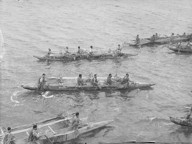 [Pacific Island men wearing leaves paddle in canoes]