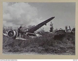 WEWAK AREA, NEW GUINEA. 1945-05-13. A SOLDIER OF 2/4 INFANTRY BATTALION SPREADING WET CLOTHES TO DRY ON A JAPANESE FIGHTER WRECKED ON THE AIRSTRIP. MASS OF HSATTERED PLANES IN THE BACKGROUND GIVES ..