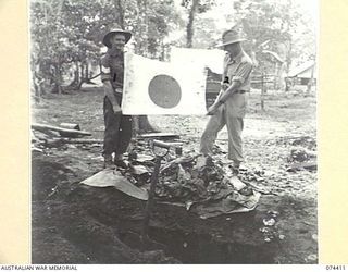 LAE, NEW GUINEA. 1944-07-01. QX30427 SERGEANT S. BROWNE (1) AND VX148000 CAPTAIN W.H.C. STRETCH, QUARTERMASTER, HEADQUARTERS, NEW GUINEA FORCE (2) DISPLAYING A JAPANESE FLAG, PORTION OF A STORE OF ..