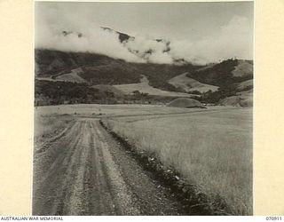 WAU - LAE ROAD, NEW GUINEA, 1944-02-26. THE ROAD RUNNING THROUGH OPEN COUNTRY THIRTY-TWO AND A HALF MILES FROM WAU, WITH THE SNAKE RIVER AND WOODED PINE TREE SLOPES ON THE RANGE IN THE BACKGROUND, ..