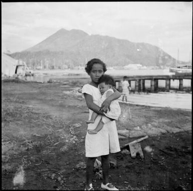 Girl holding a small child with a jetty in the background, Rabaul Harbour, New Guinea, ca. 1936 / Sarah Chinnery