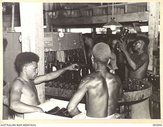 LAE, NEW GUINEA. 1944-12-12. NATIVE ASSISTANTS SUPPLIED BY ANGAU AT THE ARMY CANTEENS SERVICE SOFT DRINK FACTORY, LAE BASE SUB-AREA, REMOVING CLEAN BOTTLES FROM A BOTTLE WASHING MACHINE AND PLACING ..