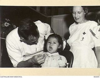 LAE AREA, NEW GUINEA. 1944-11-06. MAJOR COTTEE, 2/7 GENERAL HOSPITAL (1), ASSISTED BY PRIVATE REILLY, AUSTRALIAN ARMY MEDICAL WOMEN'S SERVICE (3), EXAMINING THE TEETH OF YENSEA, A 5 YEAR OLD ..