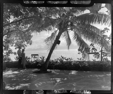 Unidentified couple at Tropique Hotel, Tahiti, standing near a palm tree looking out to the lagoon