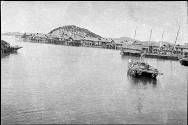 Double hull canoe in the foreground with a marine village and Port Moresby hills in the background, Papua, ca. 1923 / Sarah Chinnery