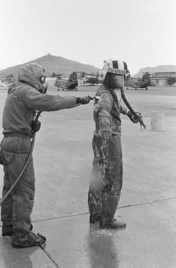 Lieutenant Colonel Thomas W. Holden, commanding officer of Marine Medium Helicopter Squadron 262 (HMM-262), undergoes decontamination procedures with the aid of Private First Class (PFC) Lipps during a combat readiness evaluation exercise. PFC Lipps is wearing nuclear-biological-chemical (NBC) gear. Visible in the background are two CH-46 Sea Knight helicopters