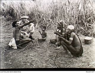 Garoka, New Guinea, 1945. Squatting on the ground, Wing Commander (Wg Cdr) Arthur Howard RAAF talks with some native Papuan porters and a child (centre). The man (left) has a gunny-sack on his back ..