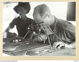 ALEXISHAFEN, NEW GUINEA. 1944-05-11. NX84716 CORPORAL S.J. HUGHES, INTELLIGENCE SECTION, HEADQUARTERS 8TH INFANTRY BRIGADE (1), EXAMINES AERIAL PHOTOGRAPHS OF THE AREA WITH A STEREOSCOPE USED TO ..