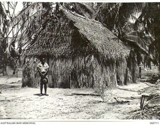 MILLINGIMBI ISLAND, NT. 1943-11-18. REVEREND KOLINIO N, SAUKURU, A FIJIAN METHODIST MISSIONARY, STANDING IN FRONT OF A FIJIAN STYLE HUT WHICH HE BUILT FOR HIMSELF