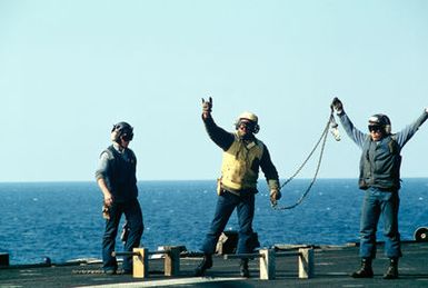 Flight deck crewmen aboard the amphibious assault ship USS GUAM (LPH 9) signal to a helicopter after setting wheel chocks into position during operations off the coast of Beirut, Lebanon