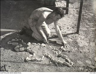 HOLLANDIA, DUTCH NEW GUINEA. 1944-06-18. A MEMBER OF NO. 24 MEDICAL CLEARING STATION RAAF WITH A MAP OF NEW GUINEA AND NEW BRITAIN MADE OF STONES AND GRAVEL IN THE DIRT SHOWING THE UNIT'S JOURNEYS