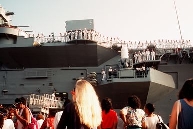 Crew members man the rails aboard the amphibious assault ship USS GUAM (LPH-9) as their relatives and friends gather on the pier for the ship's departure. The GUAM and several other U.S. Navy ships are deploying from Norfolk to the Persian Gulf in response to Iraq's invasion of Kuwait