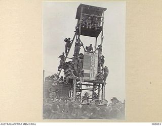 HERBERTON, QLD. 1945-01-19. HQ 9 DIVISION TROOPS CLIMB TO THE CONTROL TOWER AND SCOREBOARD TO VIEW EVENTS DURING THE 9 DIVISION GYMKHANA AND RACE MEETING HELD AT HERBERTON RACECOURSE