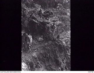 BULLDOG ROAD, NEW GUINEA. 1943-07-18. NATIVES CARRYING COMPRESSOR PARTS NEGOTIATE A NARROW AND DANGEROUS LEDGE ABOUT A MILE SOUTH OF ECCLESTON GAP. THIS LEDGE VARIES FROM EIGHTEEN INCHES TO TWO ..