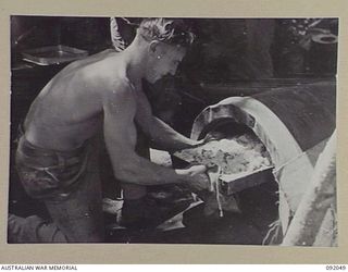 BOUGAINVILLE. 1945-05-16. CORPORAL A.R. ERBS, C COMPANY, 24 INFANTRY BATTALION, LIFTING A DISH OF BREAD AND BUTTER CUSTARD FROM AN IMPROVISED CAMP OVEN CONSTRUCTED FROM A 44 GALLON DRUM AT C ..