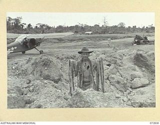 MADANG, NEW GUINEA, 1944-04-30. NX99294 SAPPER H. STAPLETON, ROYAL AUSTRALIAN ENGINEERS (1), DISPLAYING BELTS OF JAPANESE .5 INCH AMMUNITION FOUND AT MADANG AIRFIELD. A STINSON AIRCRAFT CAN BE SEEN ..