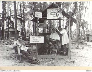BOUGAINVILLE. 1945-04-03. CORPORAL NEWTON, HEADQUARTERS 3 DIVISION (1), READING A MAGAZINE WHILE STAFF SERGEANT J.F. DAWSON, HEADQUARTERS 3 DIVISION (2), HAS HIS HAIR CUT BY PRIVATE F. NANKERVIS, ..