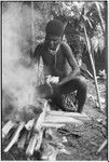 Pig festival, wig ritual, Tsembaga: adolescent boy holds plucked chicken, fire heats cooking stones for elevated oven (in background)