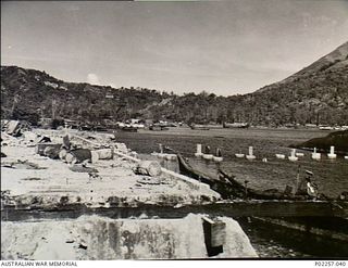 Rabaul, New Britain, 1945-10. The ruins of the Burns Philp Company's warehouse and wharves on the shores of Simpson Harbour. In the background, several barges and ships ride at anchor on the waters ..