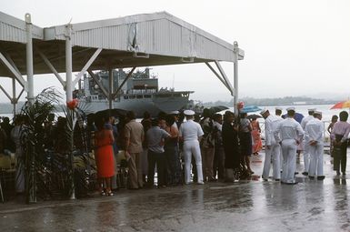 Families of the crew aboard the combat stores ship USS NIAGARA FALLS (AFS 3) stay dry under a pivilion at Naval Station Guam while welcoming the ship (background) into port