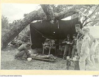 DAGUA, NEW GUINEA. 1945-03-25. NX15264 SERGEANT H.A. MARTIN (1), AMONG MEMBERS OF THE 2/2 INFANTRY BATTALION, TENDS A BATTLE CASUALTY ON A STRETCHER. THE REGIMENTAL AID POST ORDERLY VX115958 ..