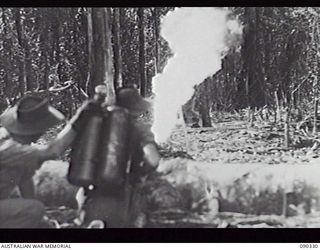 BOUGAINVILLE, 1945-04-04. A MEMBER OF THE FLAME THROWER PLATOON FROM 15TH INFANTRY BRIGADE, FIRING A MIAI PORTABLE FLAME THROWER FROM BEHIND A LOG, DURING TESTING OF FUELS, PRIOR TO USE AGAINST THE ..