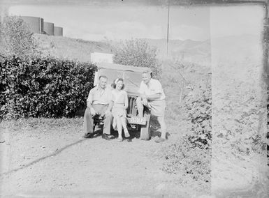 [Two men and a woman sit on front bumper of a car]