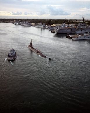 Aerial port quarter view of a "Sturgeon" class nuclear-powered submarine as it enters Pearl Harbor. The large harbor tug WAXAHACHIE (YTB 814) is visible to the left