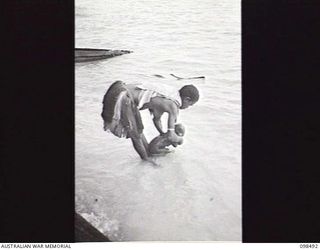 KOIL ISLAND, NEW GUINEA, 1945-10-31. A NATIVE WOMAN WASHING HER CHILD ON THE BEACH. AN OFFICIAL PARTY CONSISTING OF AN AUSTRALIAN NEW GUINEA ADMINISTRATIVE UNIT (ANGAU) DOCTOR, A RED CROSS ..