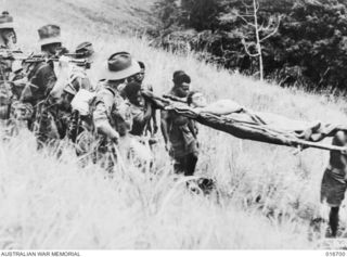 Finisterre Range, New Guinea. 1944-01. A wounded soldier is carried by local natives past a Shaggy Ridge patrol. A shot from the Department of Information film 'Jungle Patrol'