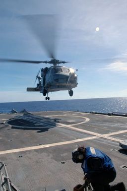 A US Navy (USN) (blue shirt) chock crew member shields himself to avoid rotor wash, as a SH-60B Sea Hawk helicopter assigned to the "Warlords" of Helicopter Anti-Submarine Squadron Light 51 (HSL-51), takes off from the flight pad aboard the USN Ticonderoga Class Guided Missile Cruiser (Aegis), USS COWPENS (CG 63) during a joint Russian Federation Navy-US Navy humanitarian assistance and disaster relief exercise conducted in the waters near the US Territory of Guam