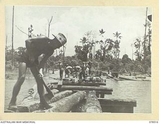 BOUGAINVILLE ISLAND, 1945-01-19. WX21039 SAPPER G.A. BOWMAN, 5TH FIELD COMPANY, TRIMMING LOGS FOR THE DECKING OF THE NEW BRIDGE ACROSS THE REINI RIVER WHILE FELLOW SAPPERS OF THE UNIT CAN BE SEEN ..