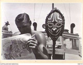 BORAM, NEW GUINEA. 1945-10-19. SERGEANT J.D. PATTERSON, 43 LANDING CRAFT COMPANY, READING THE AUSTRALIAN WOMEN'S WEEKLY. BESIDE HIM IS A MASK WHICH HE RECEIVED FROM NATIVES IN THE AREA