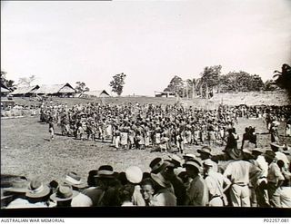 Garoka, New Guinea, 1945. Watched by a crowd of Australian soldiers (foreground), a large group of native Papuan men perform a traditional dance and 'sing-sing'. Many of the men are wearing some ..