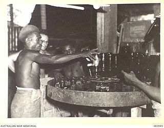 LAE, NEW GUINEA. 1944-12-12. NATIVES SUPPLIED BY ANGAU WORK UNDER THE SUPERVISION OF CORPORAL A.P. RYAN, ARMY CANTEENS SERVICE, (1), AS THEY LOAD BOTTLES INTO A CLEANING MACHINE AT A SOFT DRINK ..