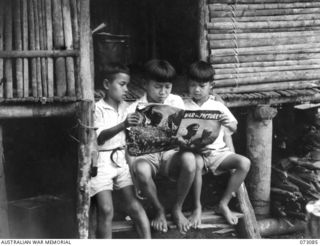 BASU RIVER VALLEY, NEW GUINEA. 1944-05-14. WAN HAN (1), JOHN WAN (2), AND JIMMY WAN (3), ON THE STEPS OF THE HOME OF WAN JIN WAH EXAMING "WAR IN PICTURES", A BRITISH PUBLICATION ON THE WAR IN ..