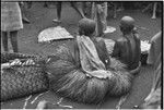 Mortuary ceremony, Omarakana: mourning women in long fiber skirts and necklaces, with heads shaved, seated beside banana leaf bundles