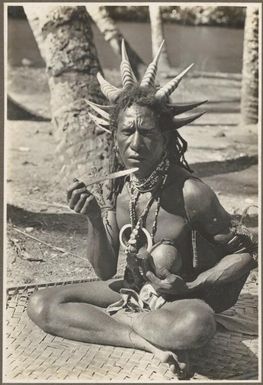 Wanigella [Wanigela] village [seated man wearing a head dress with large spikes, possibly eating betel nut] / Frank Hurley