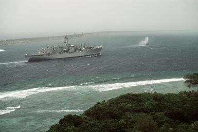 The combat stores ship USS NIAGARA FALLS (AFS 3) enters Apra Harbor while en route to Naval Station Guam. The ship is preceded by a tug spouting water. The man-made Glas Breakwater is visible in the background