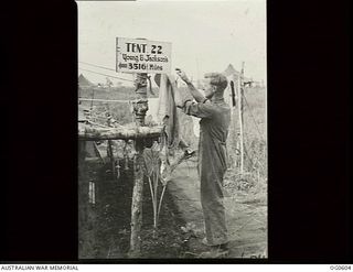 VIVIGANI, GOODENOUGH ISLAND, PAPUA. C. 1944. RAAF AIRMAN OF MILLINGTON, VIC, HANGING UP HIS WASHING NEXT TO A NOSTALGIC SIGN ON HIS TENT WHICH READS: TENT 22, YOUNG & JACKSON'S 3516 1/2 MILES