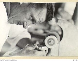 MILILAT, NEW GUINEA. 1944-08-23. QX15601 CORPORAL R.E. NORRIS, DENTAL MECHANIC, 77TH DENTAL UNIT, GRINDING ARTIFICIAL DENTURES ON A SMALL GRINDING WHEEL IN THE UNIT WORKSHOP