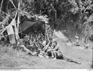 1942-10-19. THIS PICTURE, TAKEN IN THE KOKODA AREA, SHOWS A GROUP OF A.I.F. MEN IN THEIR TEMPORARY CAMP BESIDE A NATIVE "LEAN-TO" AMID SURROUNDINGS THAT CONTRAST VIVIDLY WITH THOSE THAT THEY KNEW ..