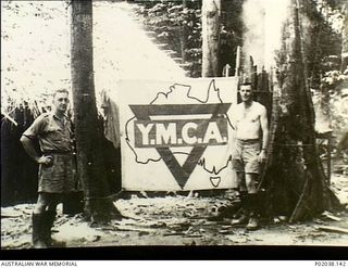 Kokoda Trail, Papua. 1942. NX239 Lieutenant Bruce Brock (left) and another soldier stand beside a Young Men's Christian Association (YMCA) banner outside a thatched hut. The hut was near the ..