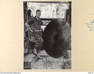 YALU AREA, NEW GUINEA, 1945-07-30. LANCE-CORPORAL J. MANTON, 2/1 FORESTRY COMPANY, ROYAL AUSTRALIAN ENGINEERS, STANDING BESIDE A BROKEN 5-FOOT CIRCULAR SAW BLADE