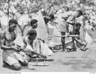 LAE, NEW GUINEA, 1944-03-25. A SERGEANT OF THE PAPUAN INFANTRY BATTALION EXAMINING TOY SAILING CANOES WHICH ARE TRADED AS NATIVE CURIOS BY RECRUITS OF THE AUSTRALIAN NEW GUINEA ADMINISTRATIVE UNIT