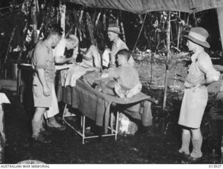 SOPUTA, PAPUA, 1942-12-23. MAIN DRESSING STATION NEAR SOPUTA. MEDICAL OFFICERS OPERATE ON A WOUNDED SOLDIER IN THE DEPTHS OF THE JUNGLE. THE OPERATING THEATRE IS IN ANKLE DEEP MUD. THE ONLY ROOF IS ..