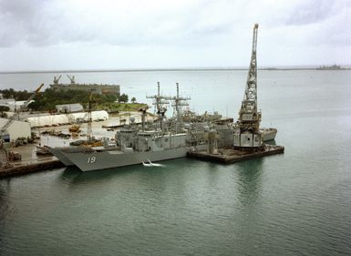 A partial port view of the guided missile frigate USS JOHN A. MOORE (FFG-19) docked at the U.S. Naval Ship Repair Facility. A large crane (YD-120-SRF) is positioned alongside the frigate