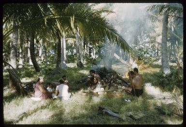 Members of the Marsters' Third Family eating a meal in a clearing among palm trees, on Palmerston Island, Cook Islands