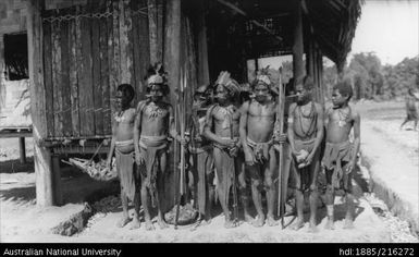 Seven men standing in front of  building, with two holding bows and arrows